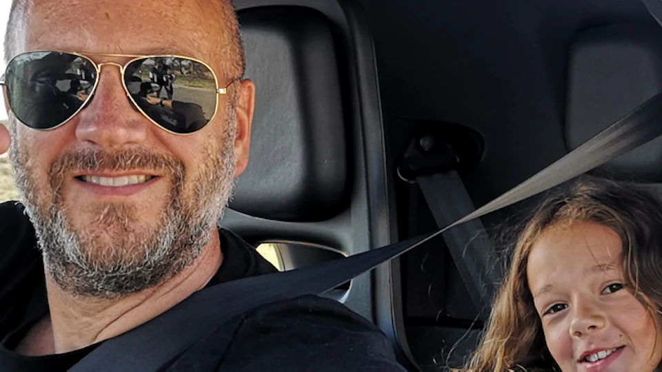 Smiling man in sunglasses and child with long hair inside a small car. The man gives a thumbs up; both wear seat belts.