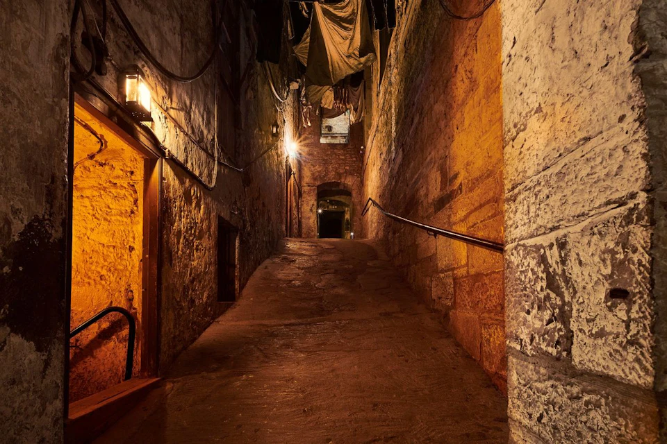Dimly lit narrow alley with stone walls, overhead clotheslines, and a sloping path leading to a dark doorway.