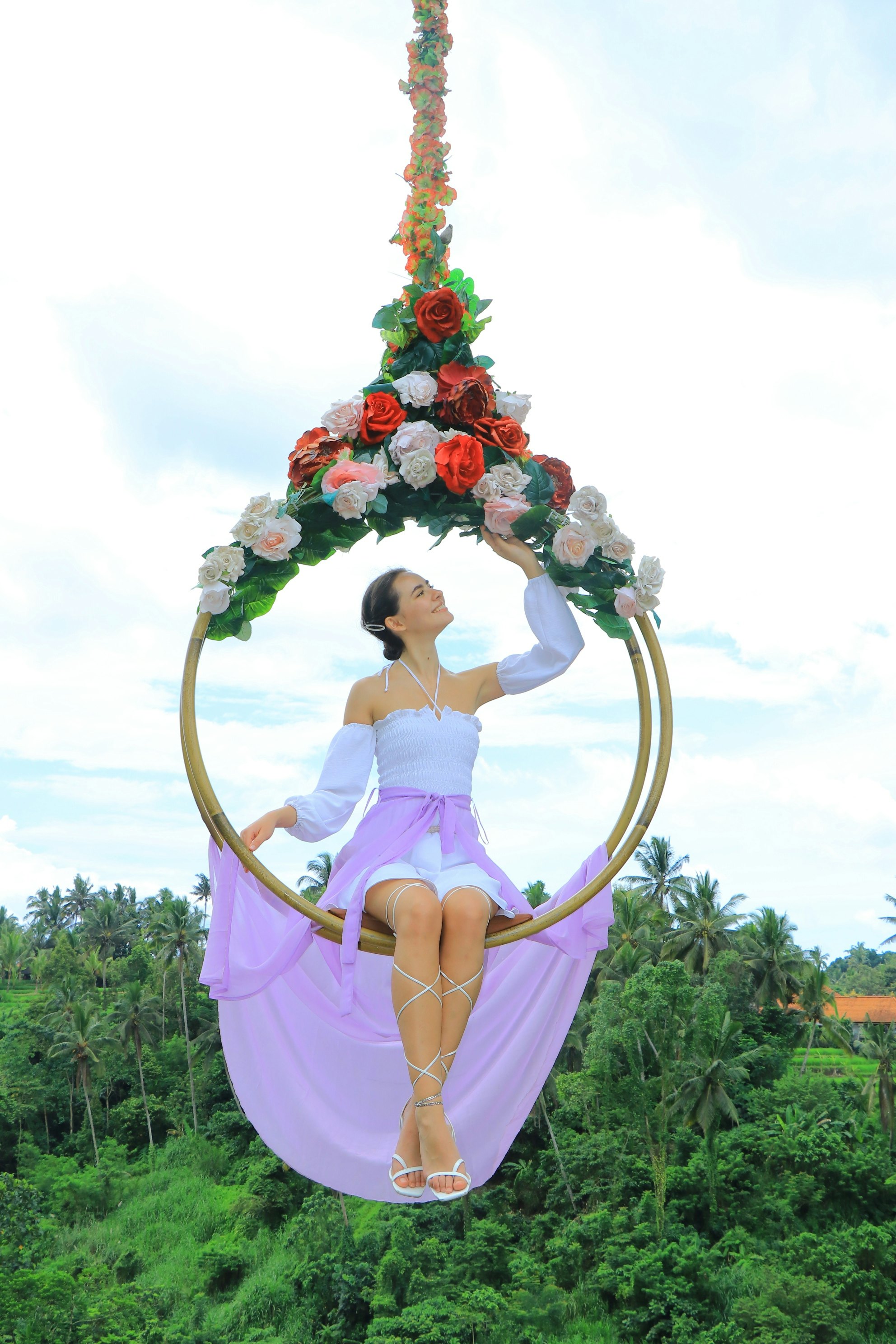 Uma mulher com um vestido branco com detalhes em roxo senta-se num baloiço adornado com rosas, rodeada por uma vegetação luxuriante.