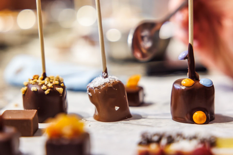 Chocolate-covered treats on sticks, decorated with nuts, coconut, and candies, with a blurred background.