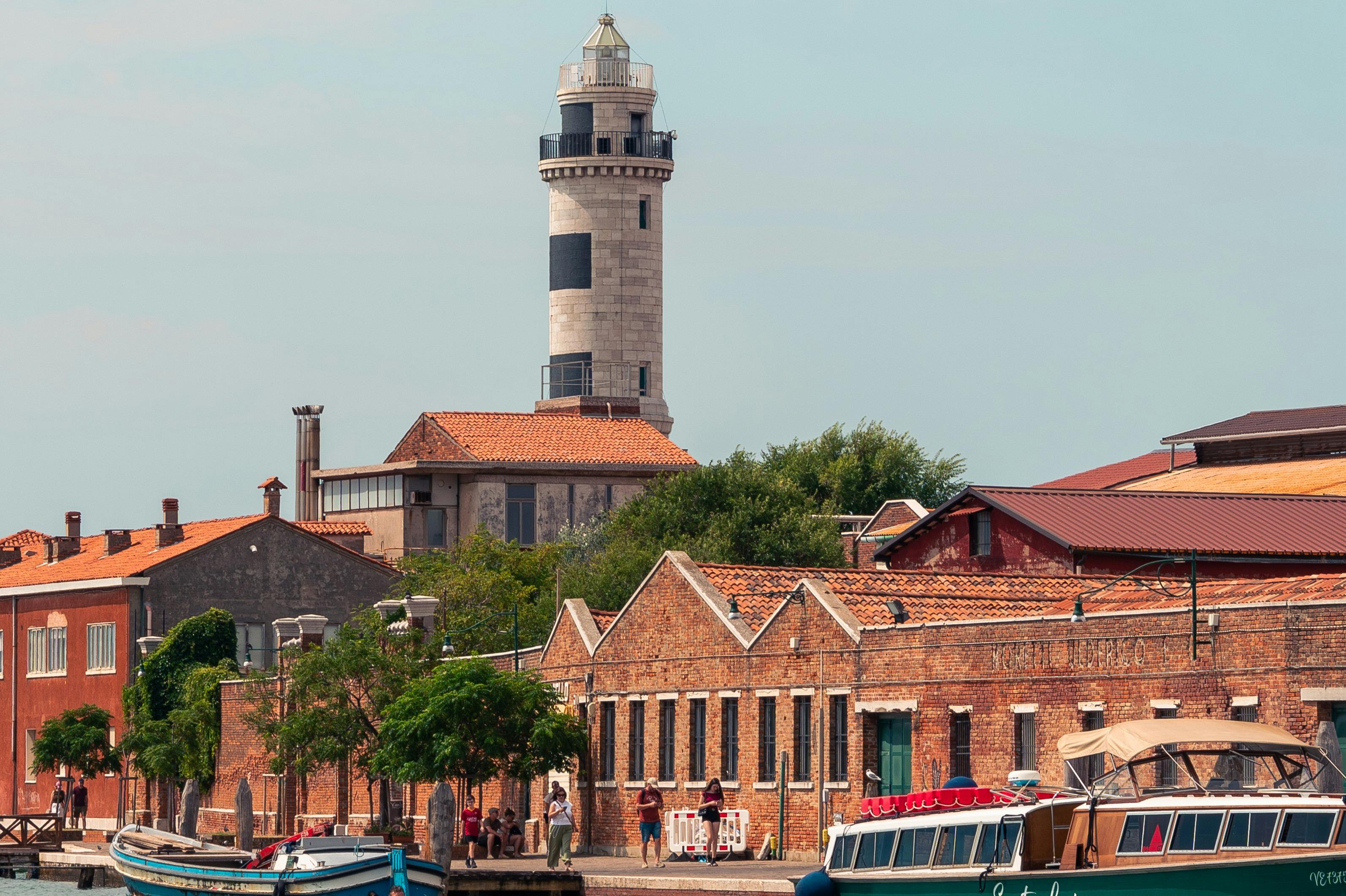 A lighthouse towers above red brick buildings and parked boats, with people walking along the waterfront on a sunny day.
