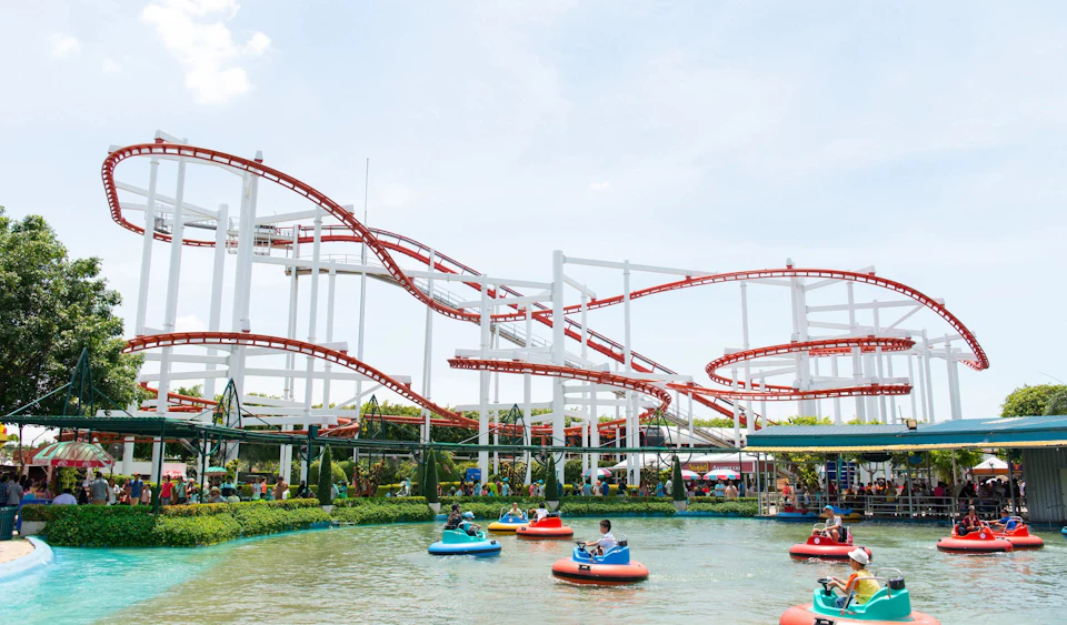 People riding colorful bumper boats in a water attraction, with a towering red and white roller coaster in the background under a blue sky.