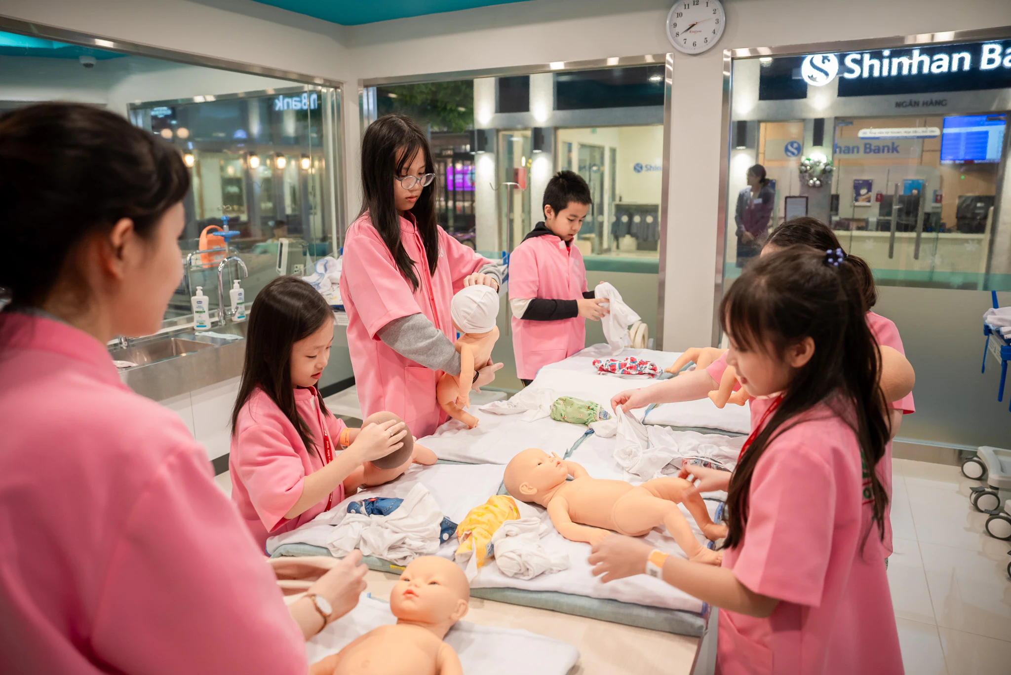 Children in pink uniforms practice caring for dolls on a table in a classroom setting.