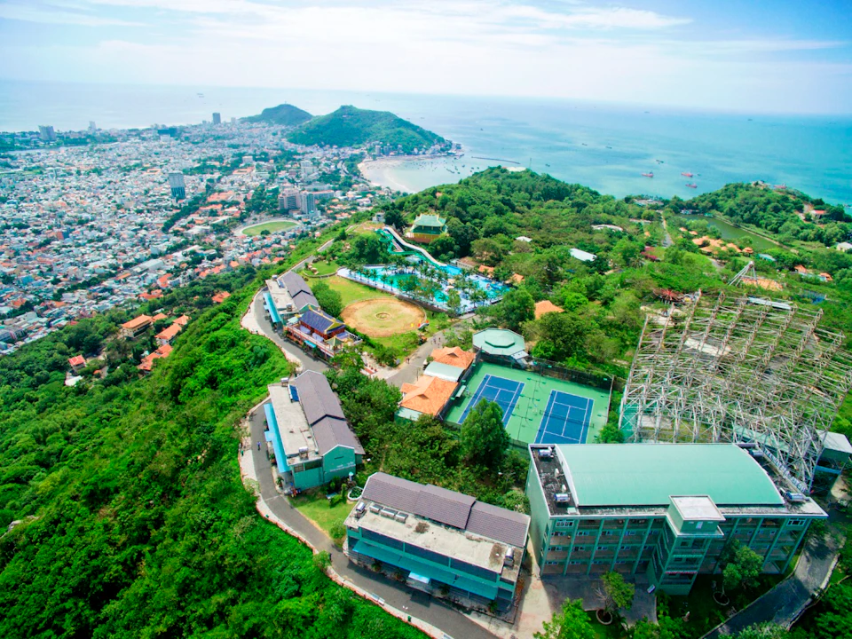Aerial view of a coastal city with green hills, buildings, tennis courts, and a sea in the background under a blue sky.