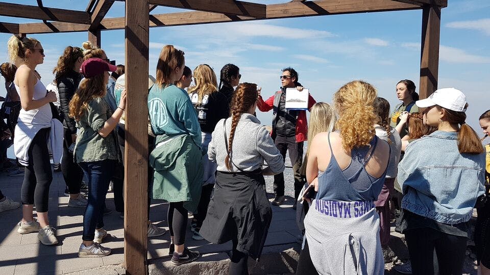 Un gruppo di persone riunite intorno a una persona con un foglio in mano che sembra tenere un discorso o una presentazione all'aperto.