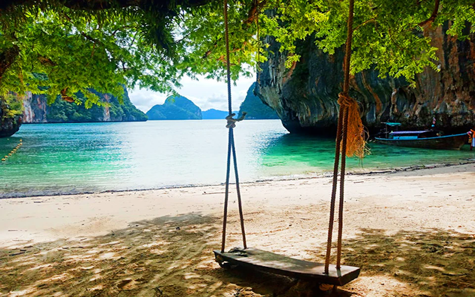 A wooden swing hanging from a tree on a sandy beach overlooking clear blue water and limestone cliffs in the background.