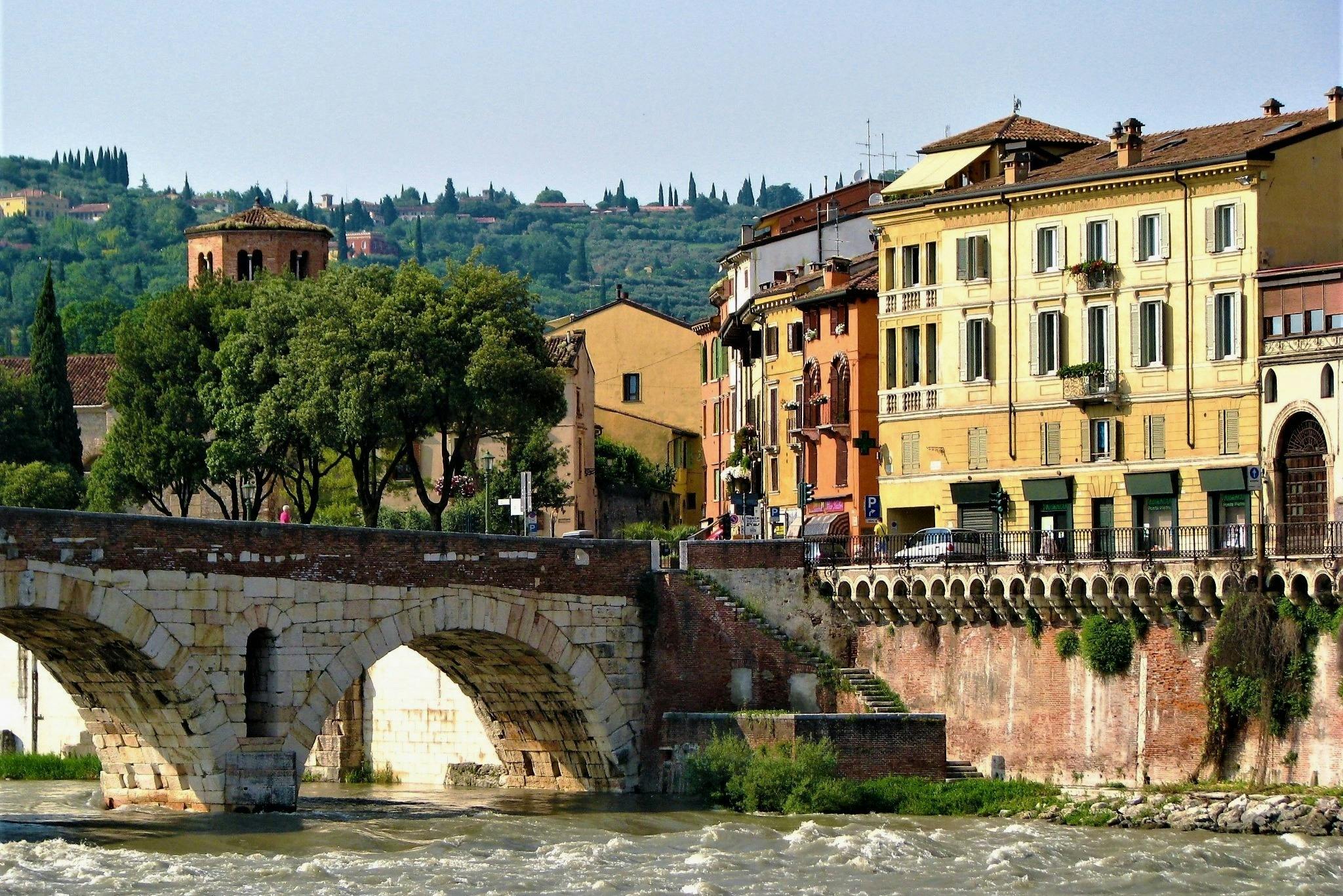 A historic stone bridge over a river with colorful buildings and trees in the background, under a clear sky.