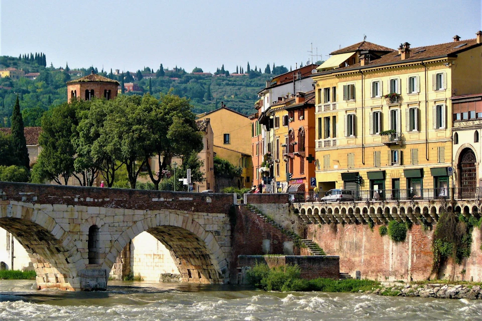 A historic stone bridge over a river with colorful buildings and trees in the background, under a clear sky.