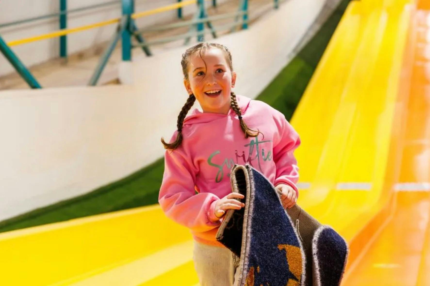 A smiling child wearing a pink hoodie holds a mat, standing by a yellow slide in an amusement park.