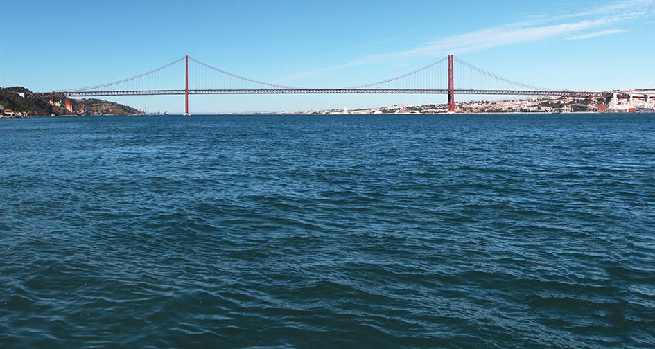 A red suspension bridge spans across a wide body of blue water, with a city visible in the background under a clear sky.