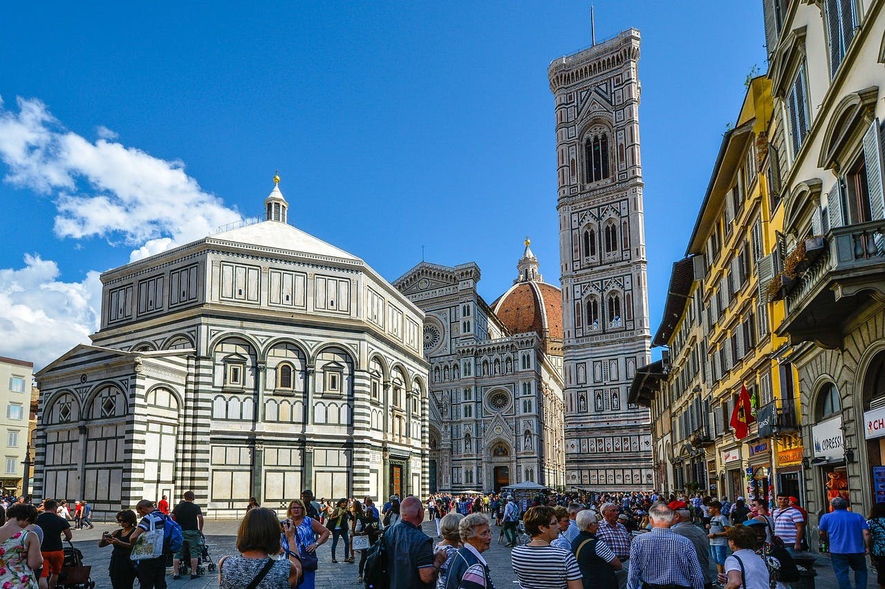 A busy square in front of the Florence Cathedral, featuring the Baptistery and Giotto's Campanile, with many people walking and taking photos.