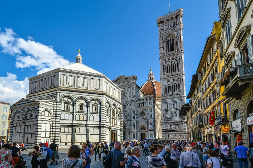 A busy square in front of the Florence Cathedral, featuring the Baptistery and Giotto's Campanile, with many people walking and taking photos.