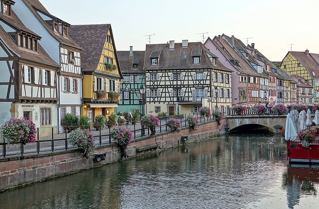 Una fila di case a graticcio colorate lungo un canale con fioriere e un piccolo ponte pedonale.