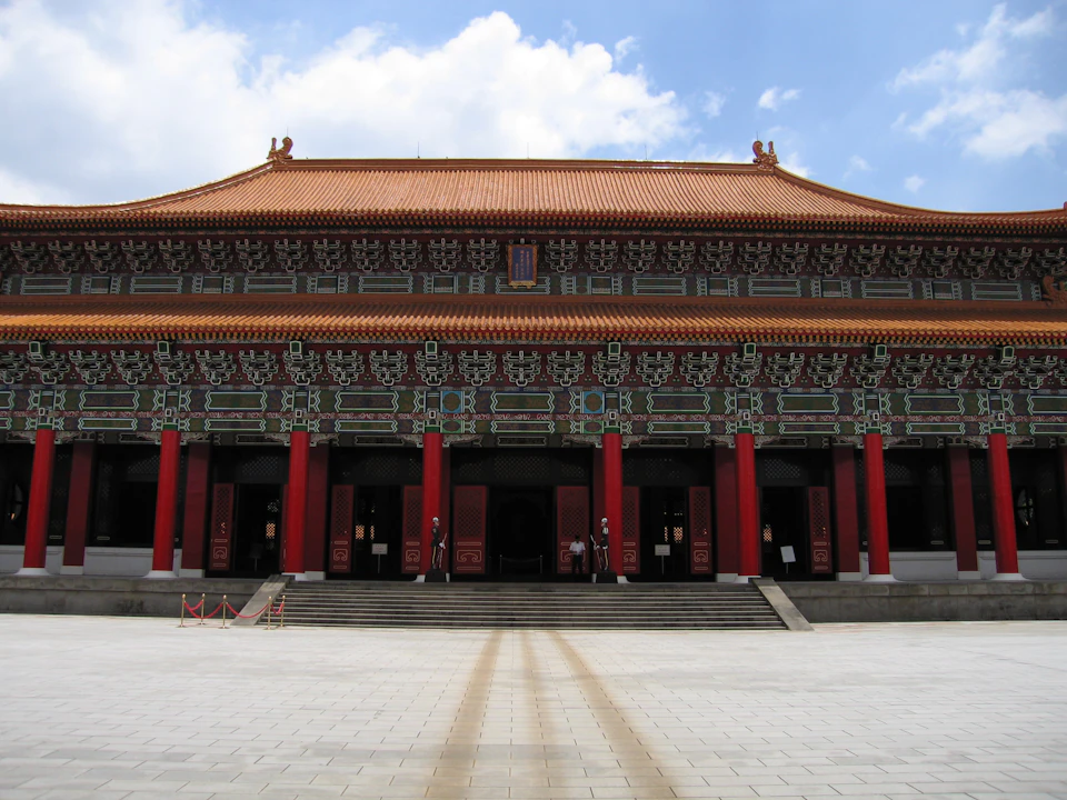 Large traditional building with ornate details, red columns, and a tiled roof. Wide steps lead to the entrance. Clear sky above.