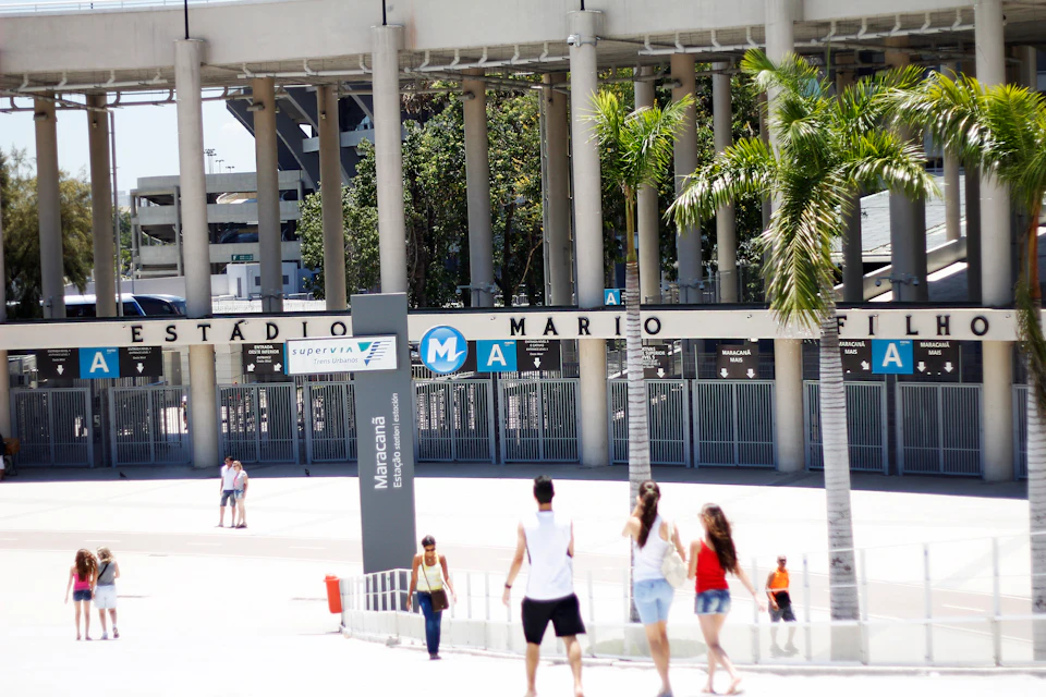 People walking outside Maracanã Stadium in daylight, with palm trees and the stadium entrance visible in the background.