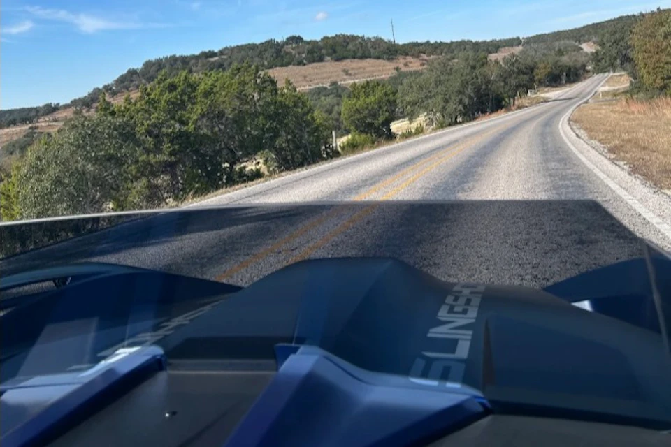 View from a vehicle's cockpit on a rural road surrounded by trees and hills under a clear sky.