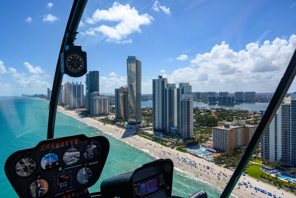 View from a helicopter cockpit overlooking a cityscape with tall buildings, a beach, and turquoise ocean under a blue sky.