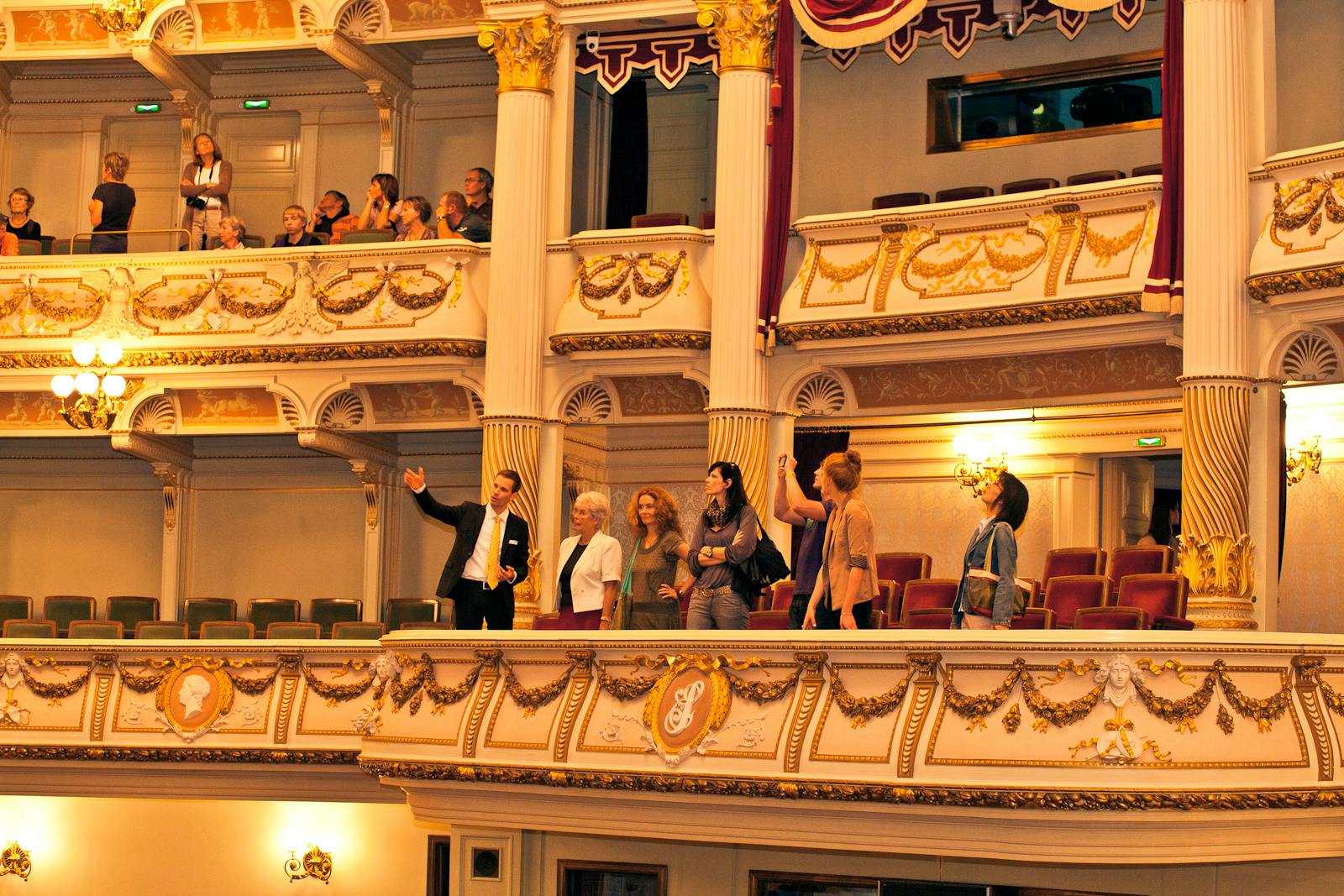 People standing on an ornate theater balcony, admiring the view. Upper balconies have additional spectators.