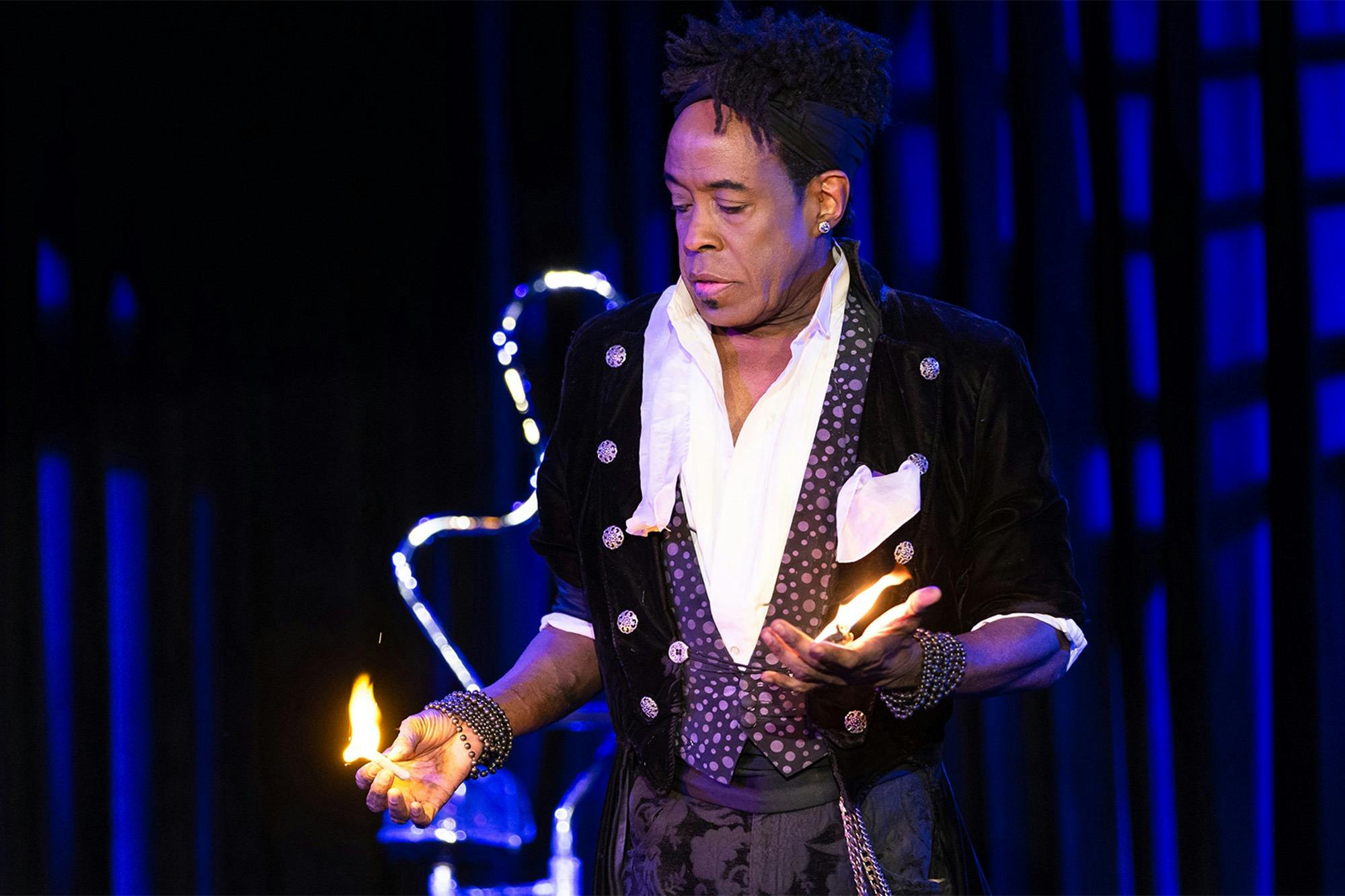 A performer in an elaborate outfit holding fire in both hands on a dark stage with blue lighting.