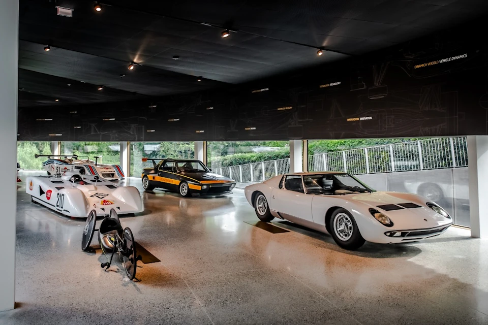 A sleek exhibition space displays a white racing car, a yellow and black vintage car, and a white sports car.