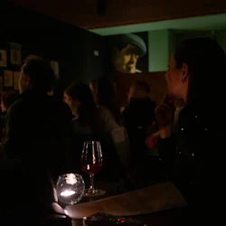 Människor sitter i ett svagt upplyst rum, några håller drinkar, med inramade bilder på väggen och ett ljus på ett bord.