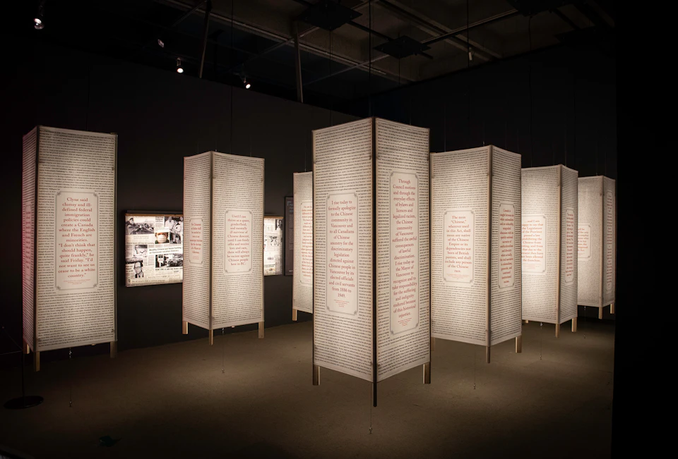 Exhibit with hanging panels of text in a dimly lit room; illuminated boards with historical photos are visible in the background.