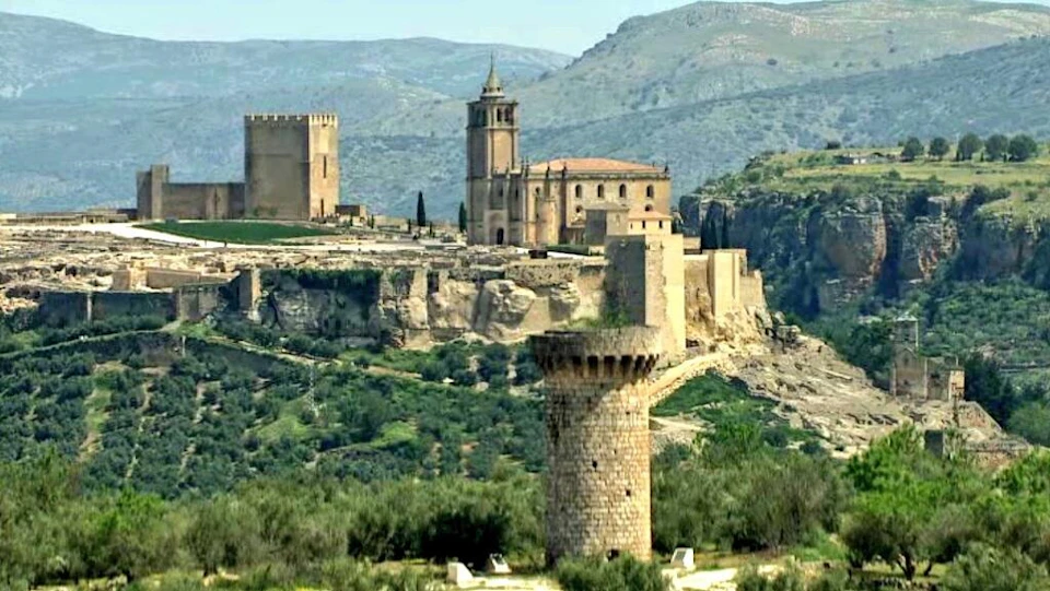 A historic stone castle and tower sit atop a rugged hill, surrounded by green landscape and distant mountains under a clear sky.