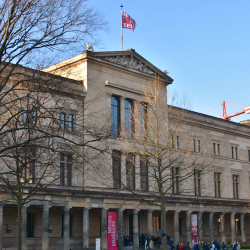 Stor, neoklassisk byggnad med pelare, en "Neues Museum" -banner och människor samlades utanför. Lövlösa träd och en röd kran syns.