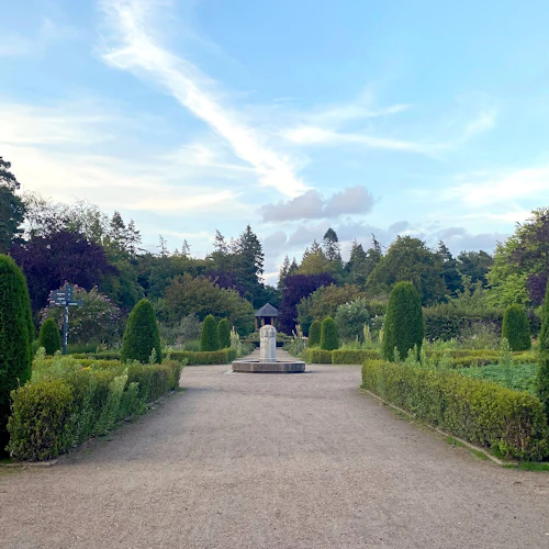 En anlagd trädgård med symmetriska häckar, höga trimmade träd och en central fontän under en delvis molnig blå himmel.