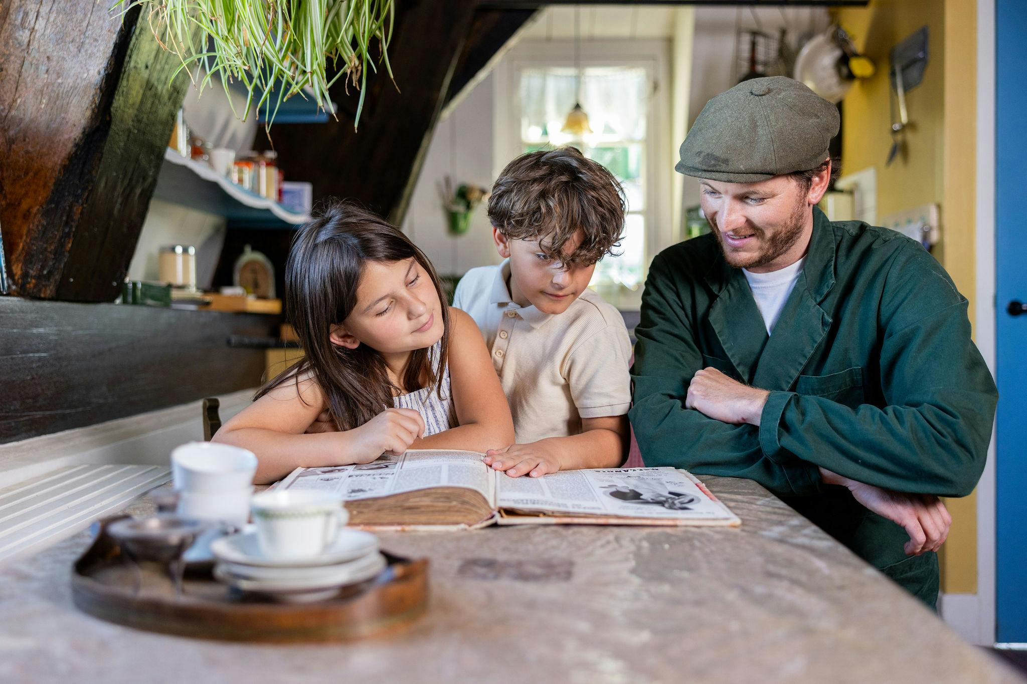 Muž a dvě děti sedí u stolu a dívají se na otevřenou knihu v útulné kuchyni.