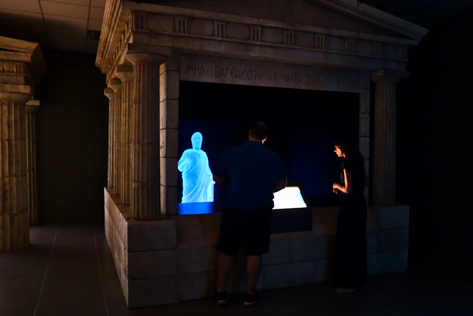 Two people observe a blue holographic statue inside a stone structure with ancient Greek architectural details.