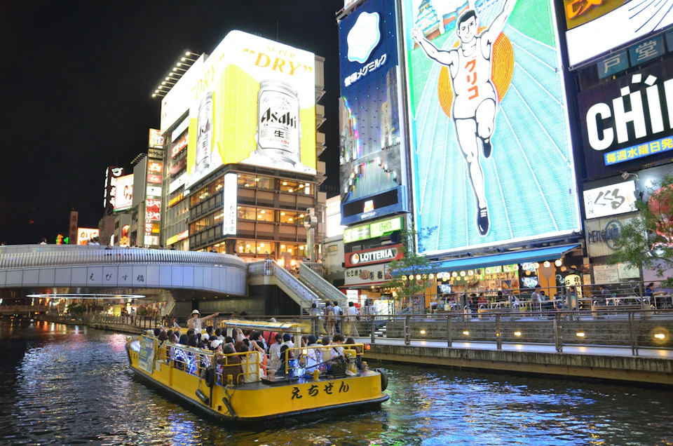 A brightly lit urban canal scene at night with a tour boat and colorful illuminated advertisements, including a famous runner sign.