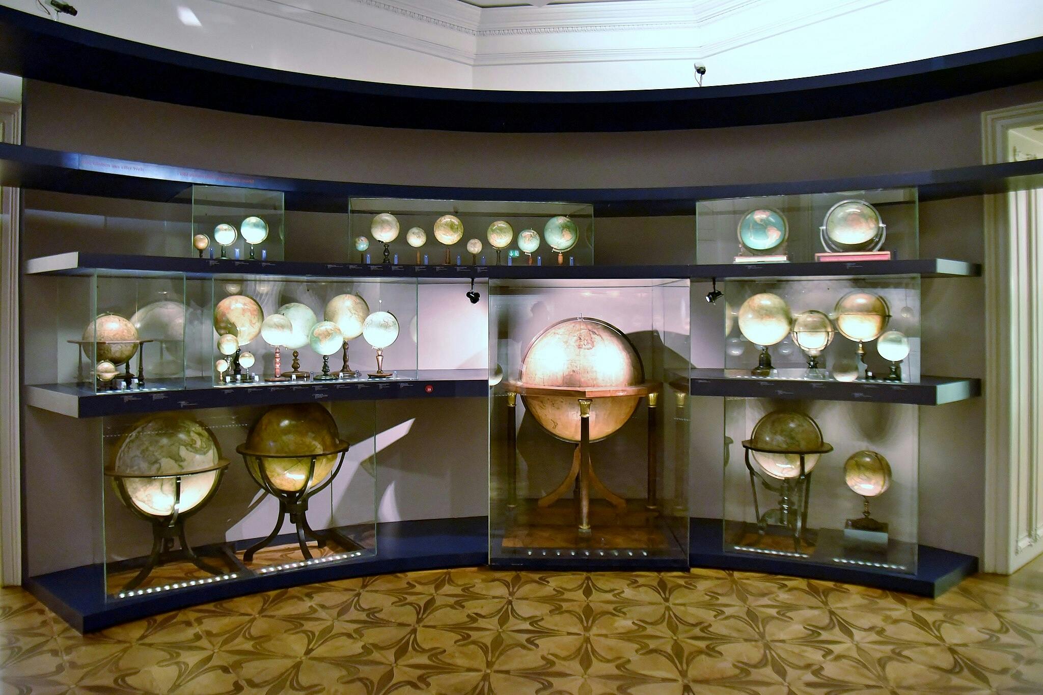 在架子上靠墙布置的玻璃盒中展示不同尺寸和年龄的各种地球仪。