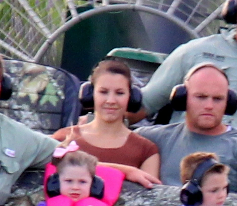 Six people, including a young girl and boy, ride on an airboat through grassy wetlands. The propeller and greenery are visible.