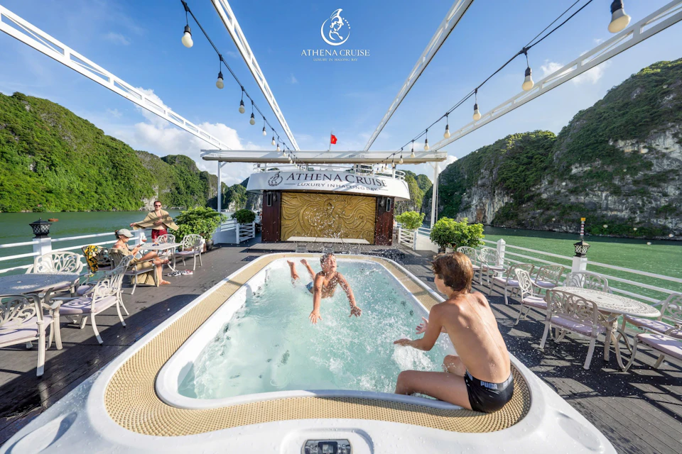 Two people enjoying a small pool on a luxury cruise ship deck with scenic cliffs in the background under a clear blue sky.