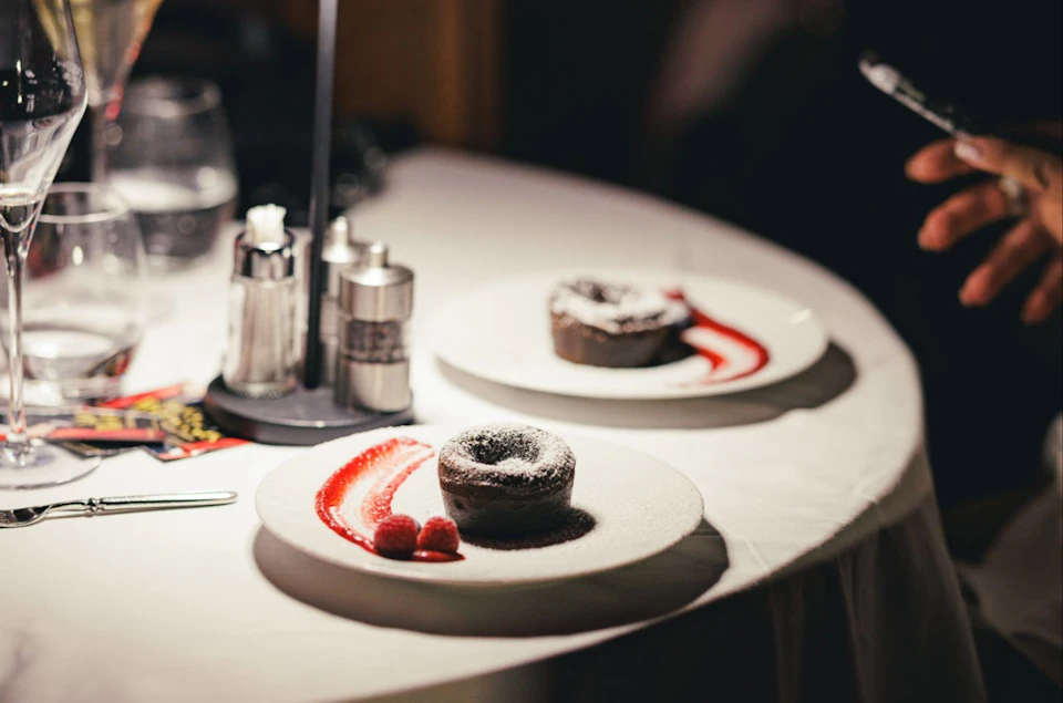 Two plates with chocolate desserts, garnished with a red sauce and raspberries, on a white tablecloth.