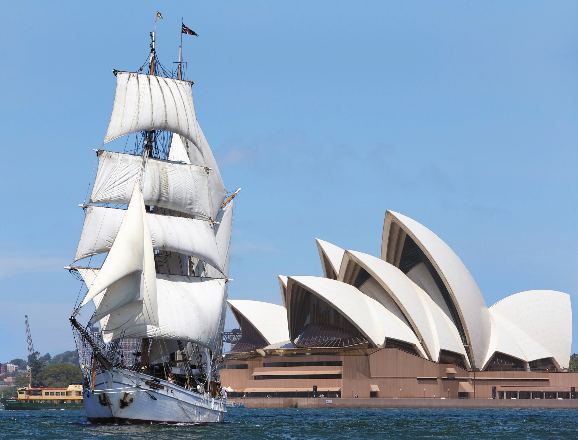 A sailing ship with white sails is in the water near the Sydney Opera House under a clear blue sky.