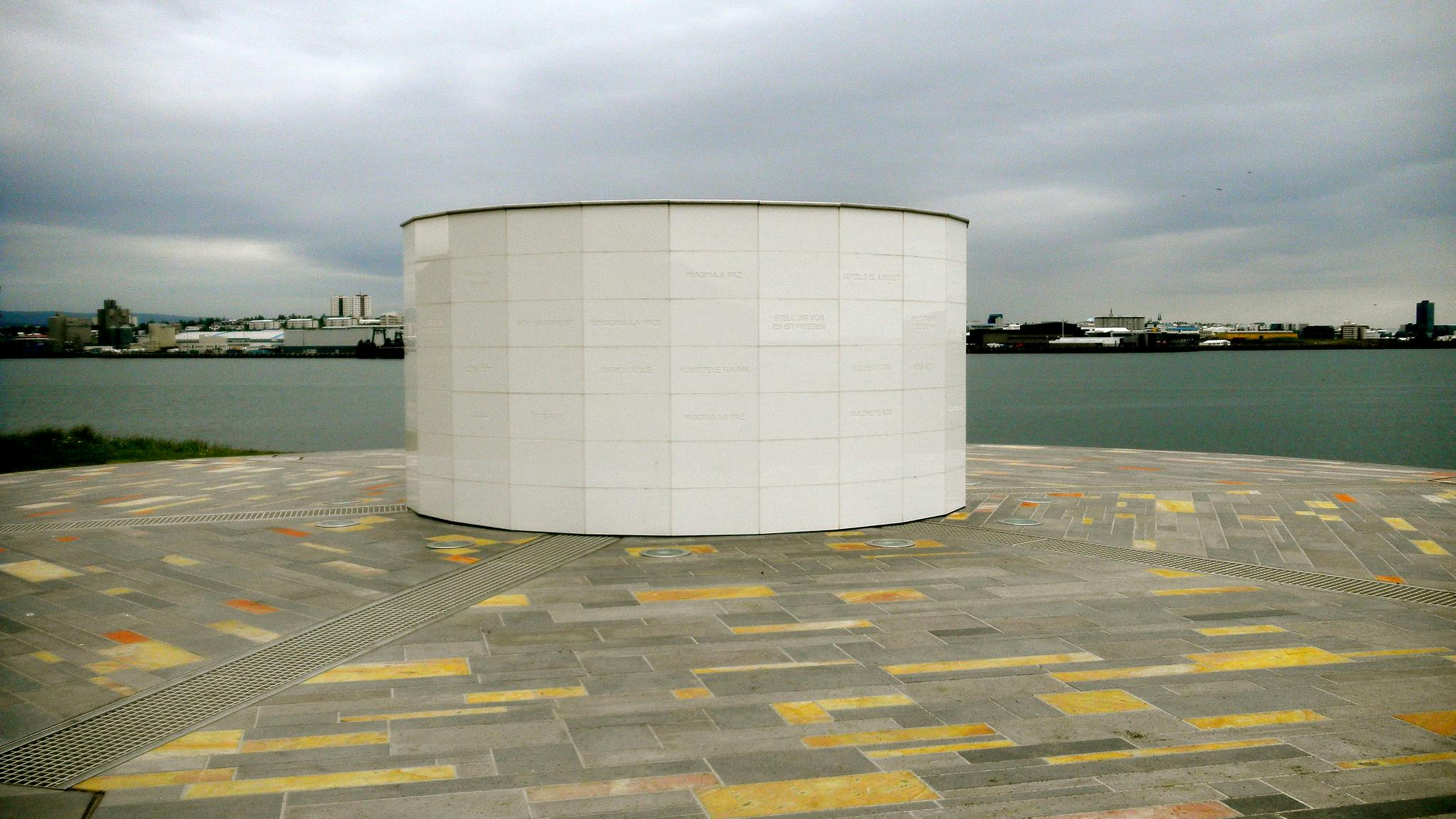 A cylindrical white structure on a patterned pavement by a body of water, with an urban skyline in the background under a cloudy sky.
