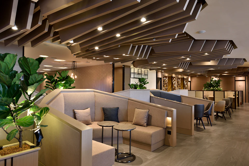Modern lounge with beige seating, small round tables, indoor plants, and a geometric ceiling design.