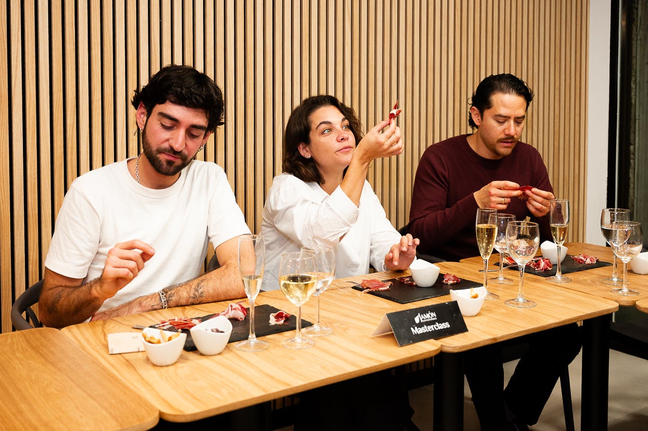 Two people sit at a wooden table with food and drinks. One observes their hand, while the other examines a small piece of food.