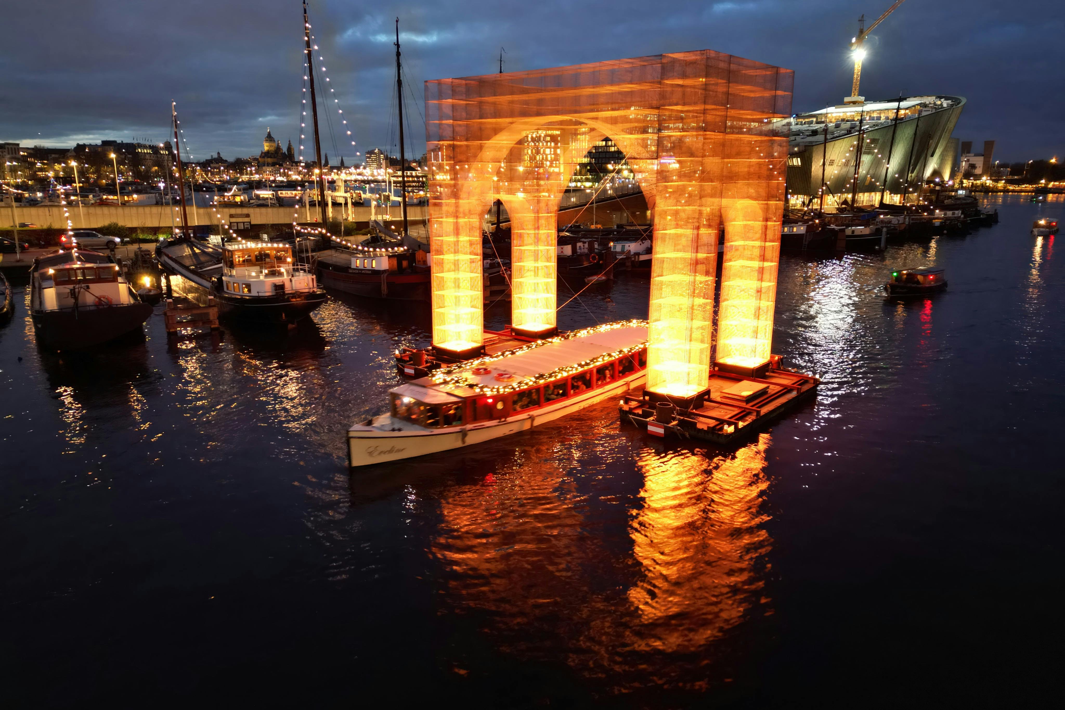 Beleuchtete Bogenkonstruktion auf dem Wasser mit einem Boot darunter, umgeben von anderen Booten und Stadtlichtern in der Abenddämmerung.