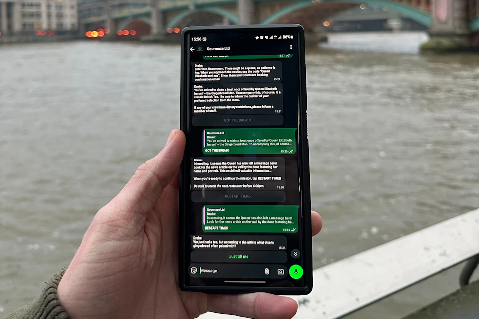 A hand holding a smartphone displaying a messaging app with a conversation visible. A river and bridge are in the background.