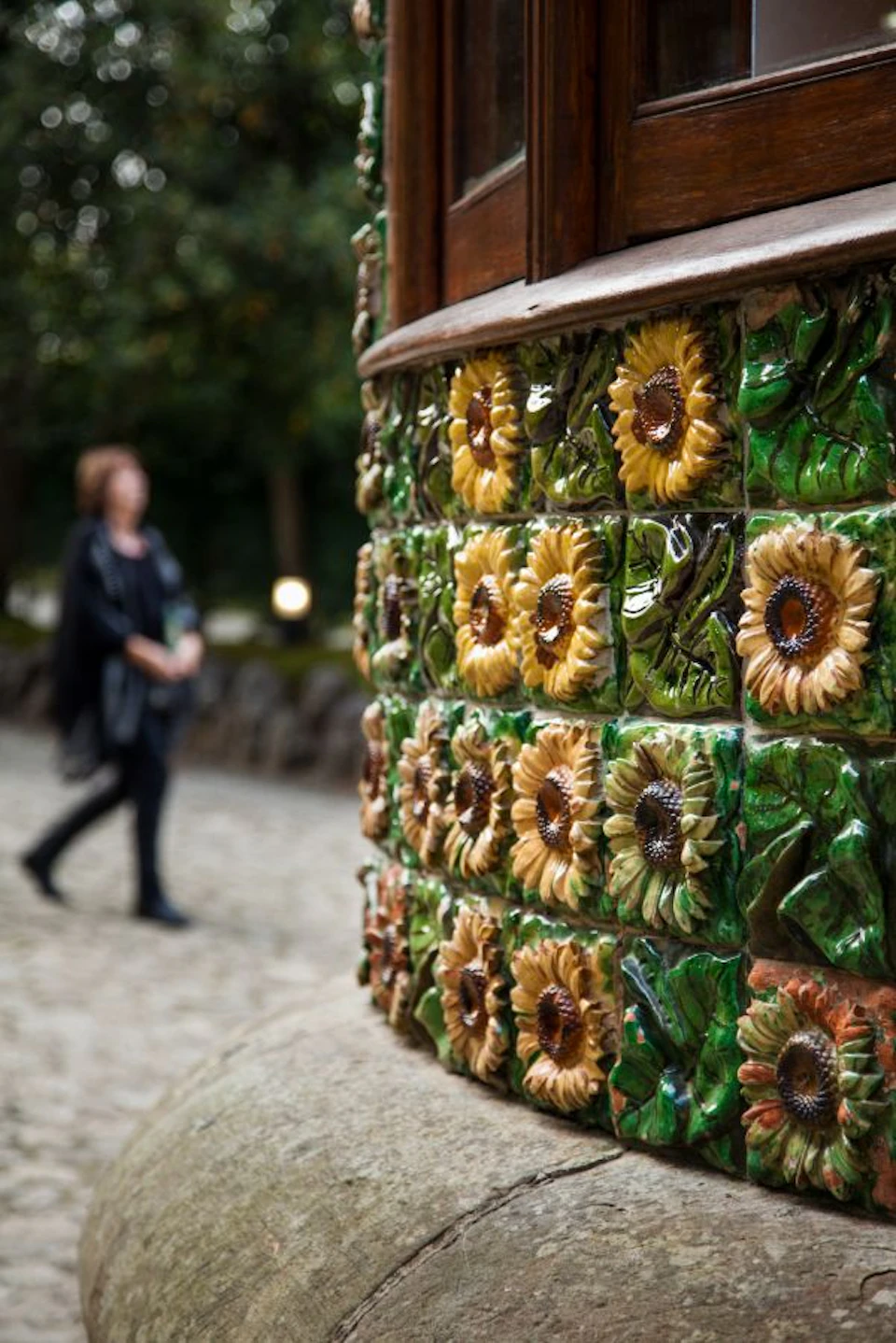 Close-up of a wall adorned with ceramic sunflower tiles. A person in dark clothing is blurred in the background.