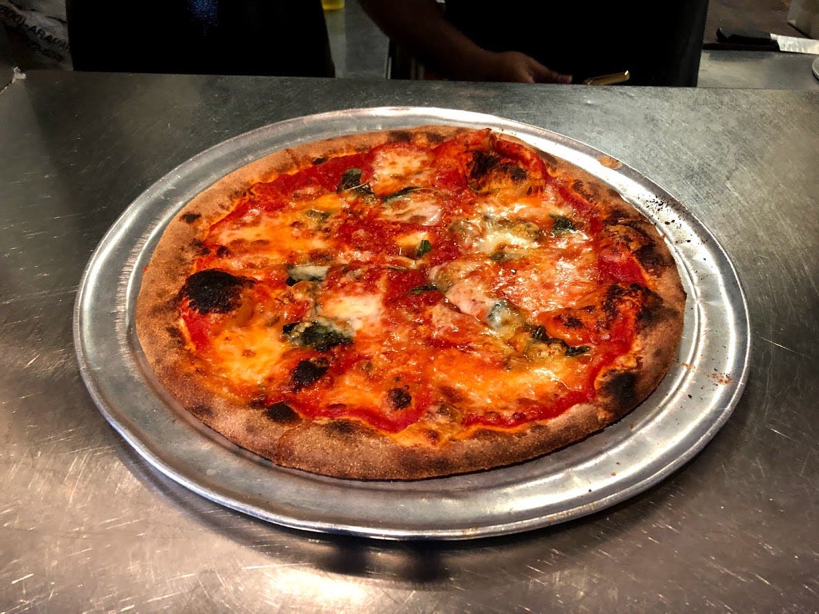 A cooked margherita pizza on a metal tray, with visible charred edges and melted cheese on a countertop.