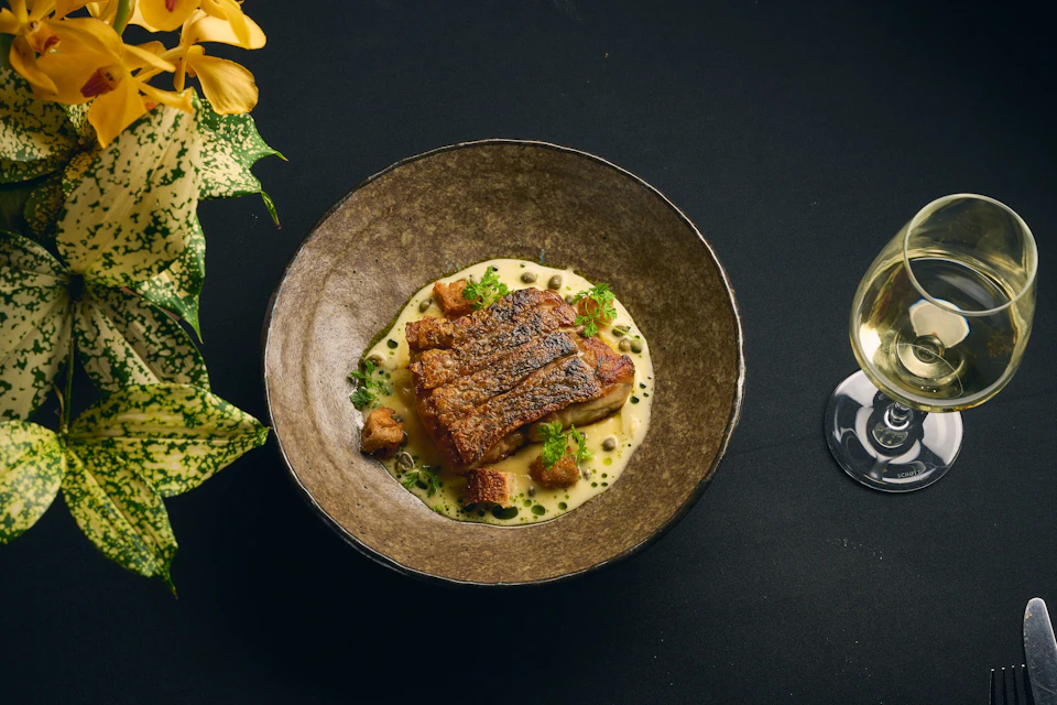 A plated dish of crispy-skinned fish on a creamy sauce with herbs and croutons, set on a black surface near a glass of wine.