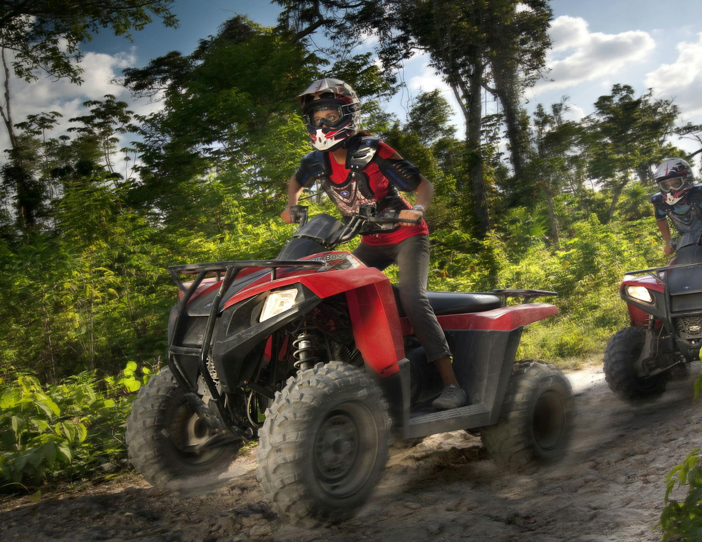 Ένα άτομο με προστατευτικό εξοπλισμό οδηγεί ένα κόκκινο ATV σε ένα χωματόδρομο μέσα σε ένα καταπράσινο δάσος.