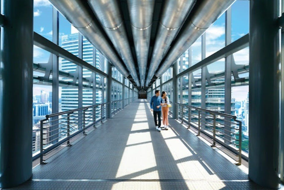 Una pareja caminando por un moderno puente elevado de cristal con un diseño industrial y edificios de la ciudad visibles en el exterior.