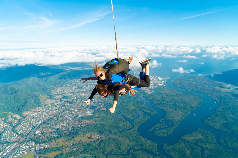 Two people tandem skydiving above a scenic landscape with forests, a city, and bodies of water below, under a clear blue sky.
