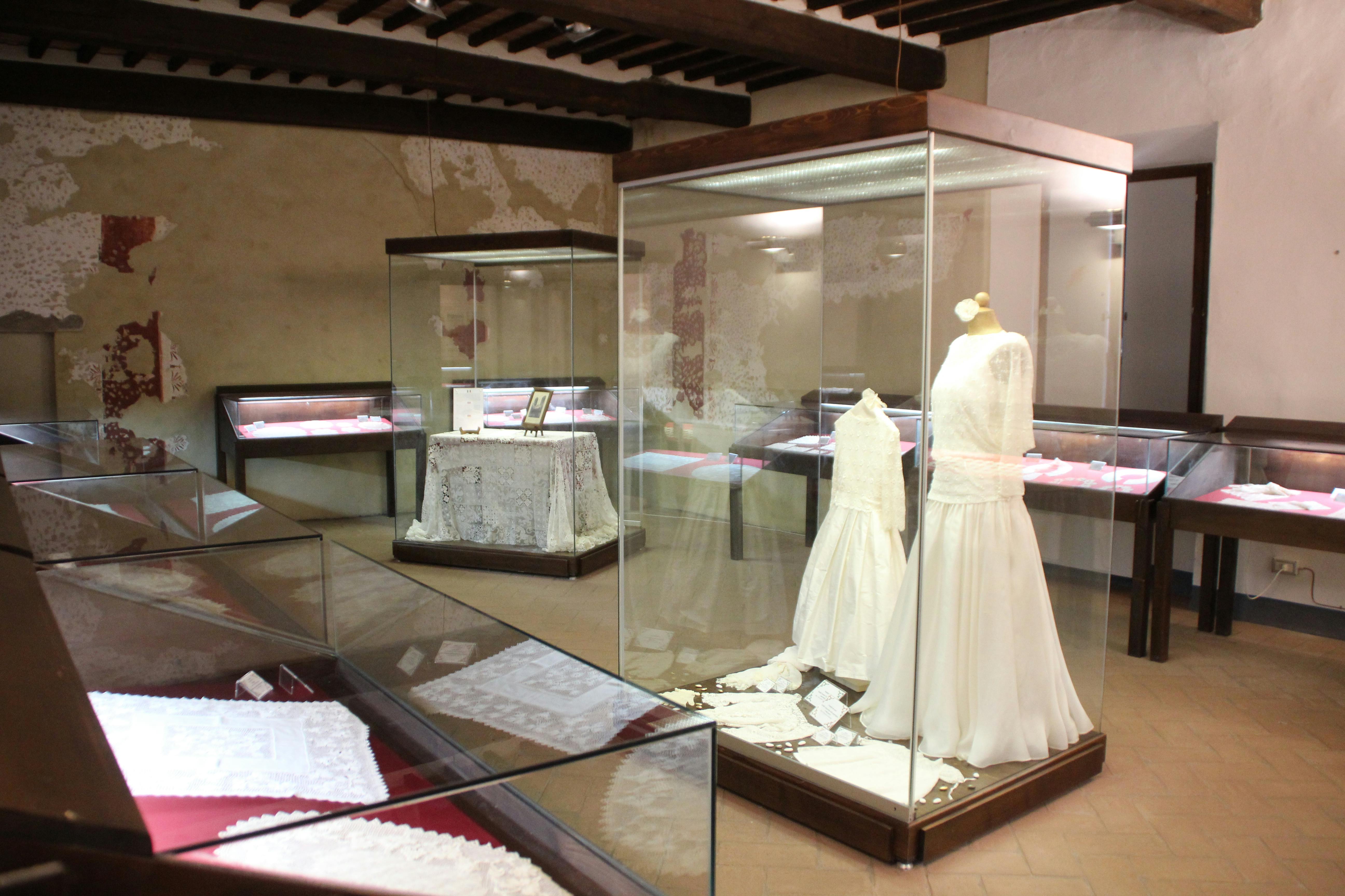 웨딩 드레스, 레이스 패브릭, 오래된 사진, 전통 직물이 전시된 유리 케이스가 있는 전시실입니다.