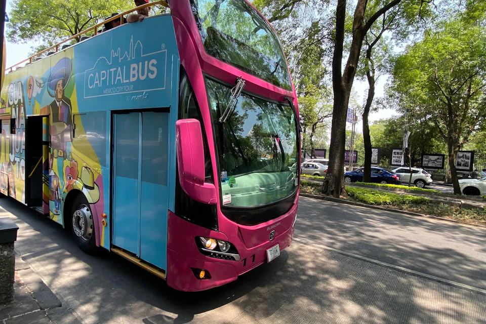 A double-decker tour bus labeled "Capital Bus" in Mexico City is parked on a tree-lined street.